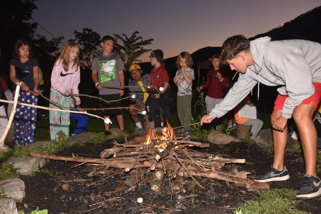 Campers and staff member roasting marshmallows around campfire at night