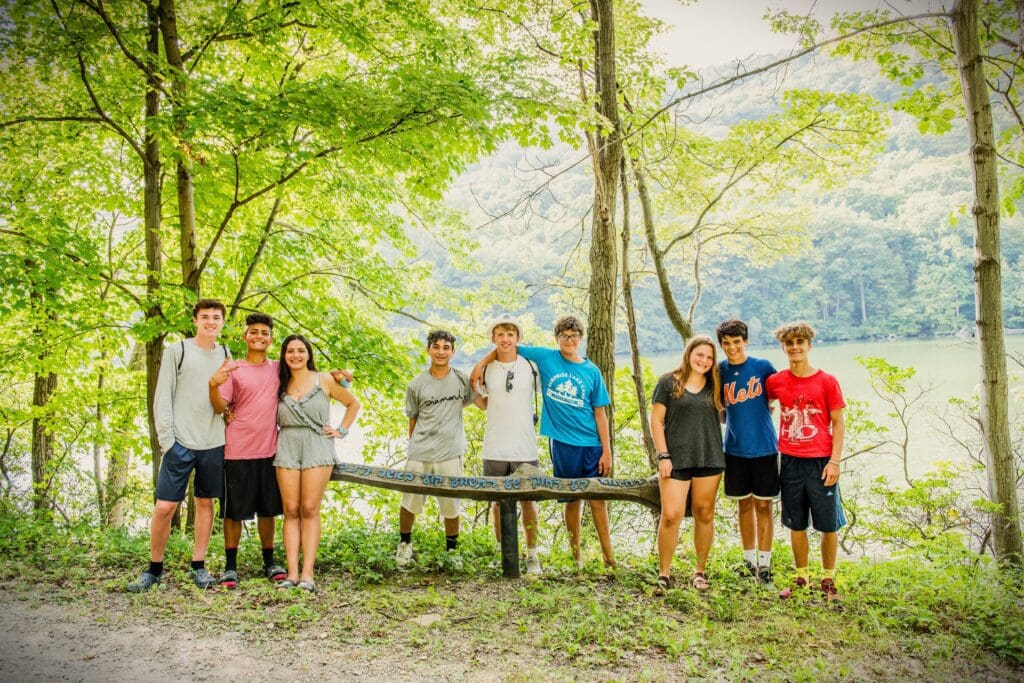 Teen SLC campers pose inn the woods, next to a wooden bench.
